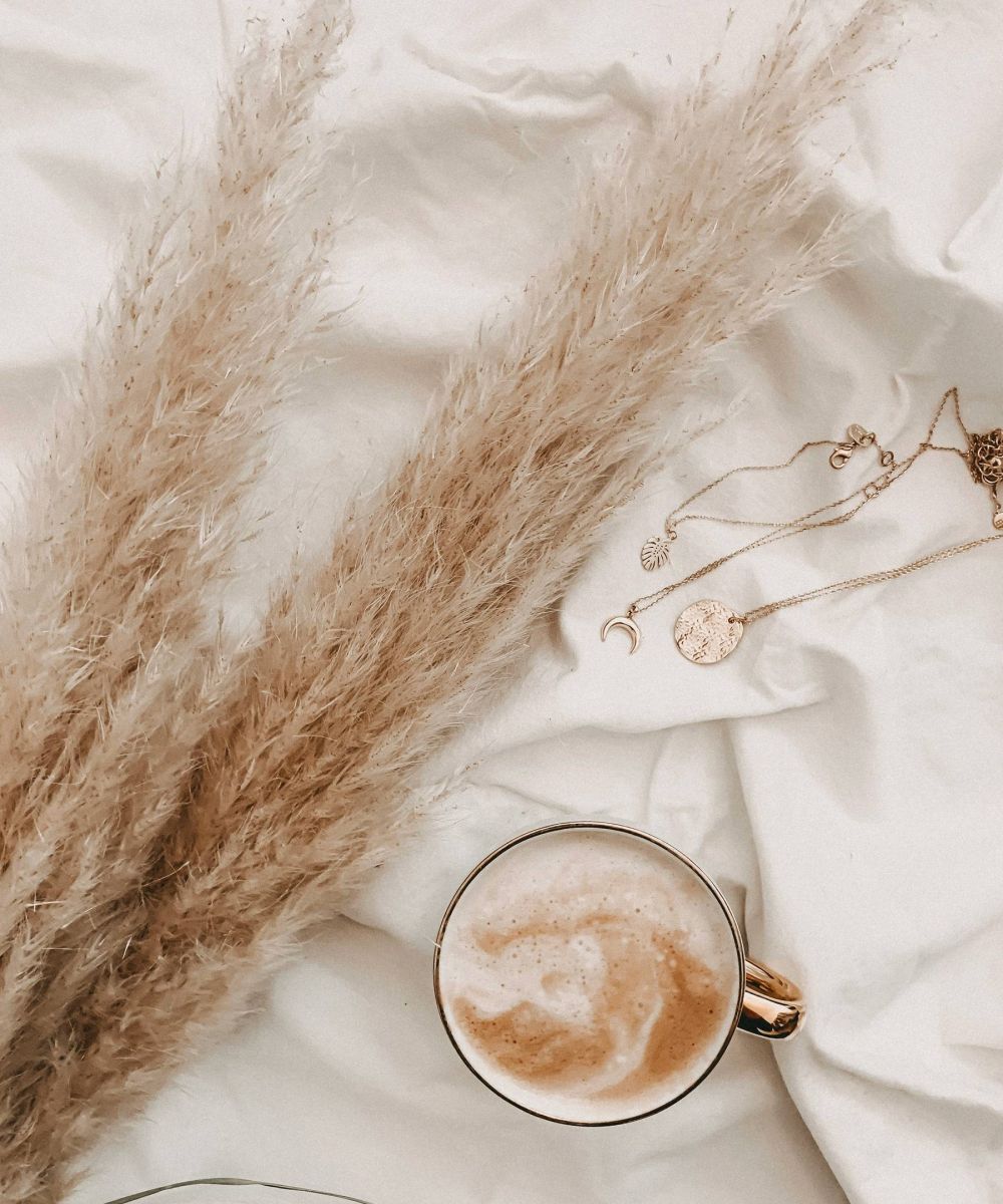 A cup of coffee, gold necklaces, and pampas grass on white fabric.